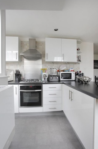 Wide view of the kitchen — gloss-white cabinets, gas hob, stainless extractor, microwave on the counter, with grey-tile floor visible at the threshold.